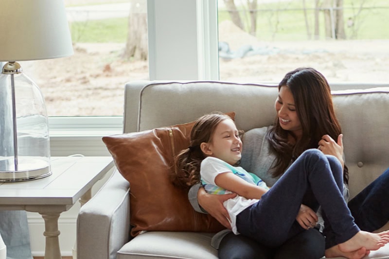 How Can I Make My Furnace Last Longer? Photo of a mother and daughter laughing and playing together on a couch in their living room.